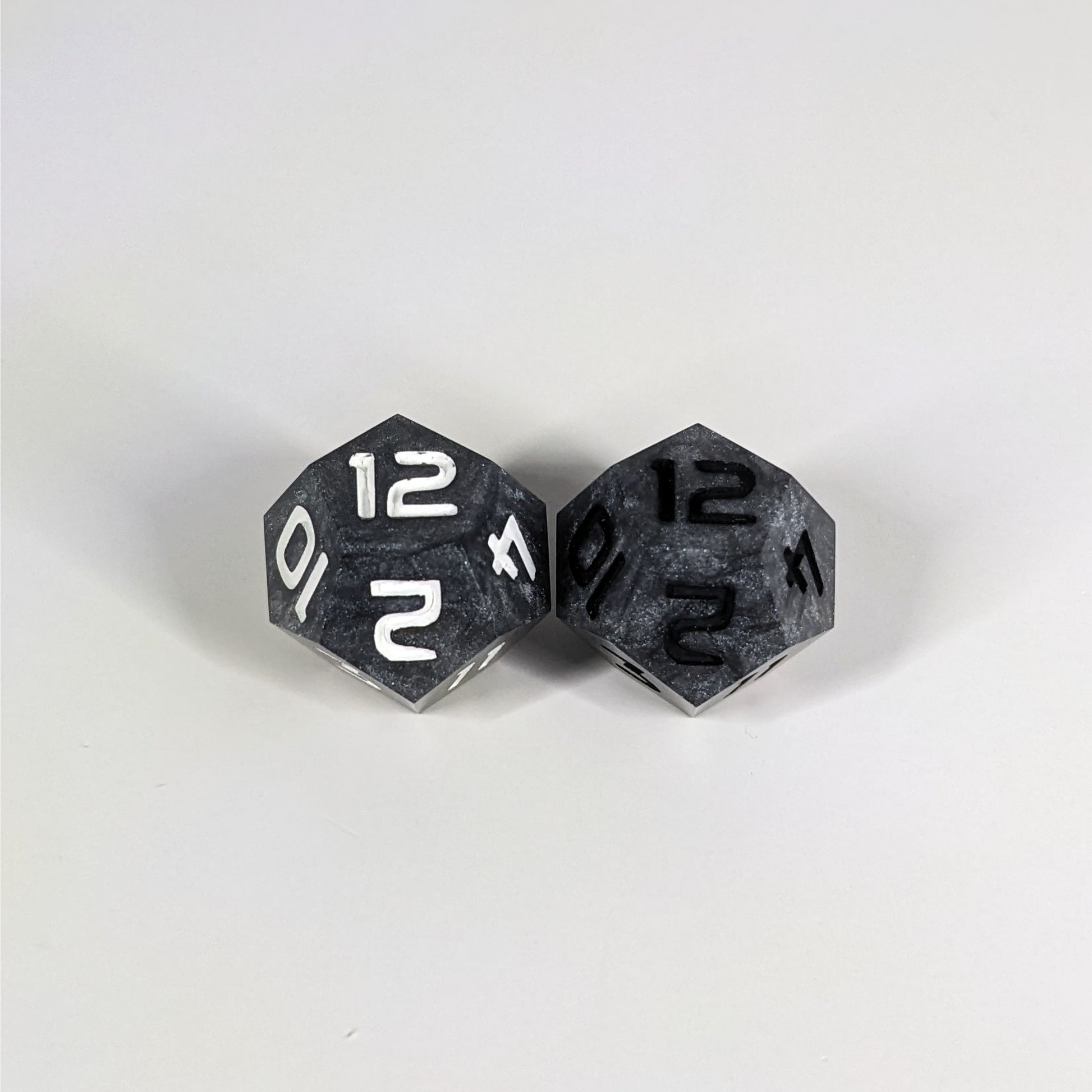 Two handmade d12s sit with the 12 faces up on a white background. The dice are dark silver, and while they also have a touch of gold the gold is not visible at this angle. The die on the left has white numbers, the die on the right has black numbers.