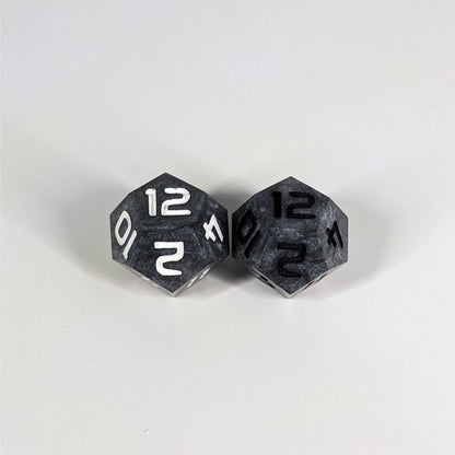 Two handmade d12s sit with the 12 faces up on a white background. The dice are dark silver, and while they also have a touch of gold the gold is not visible at this angle. The die on the left has white numbers, the die on the right has black numbers.