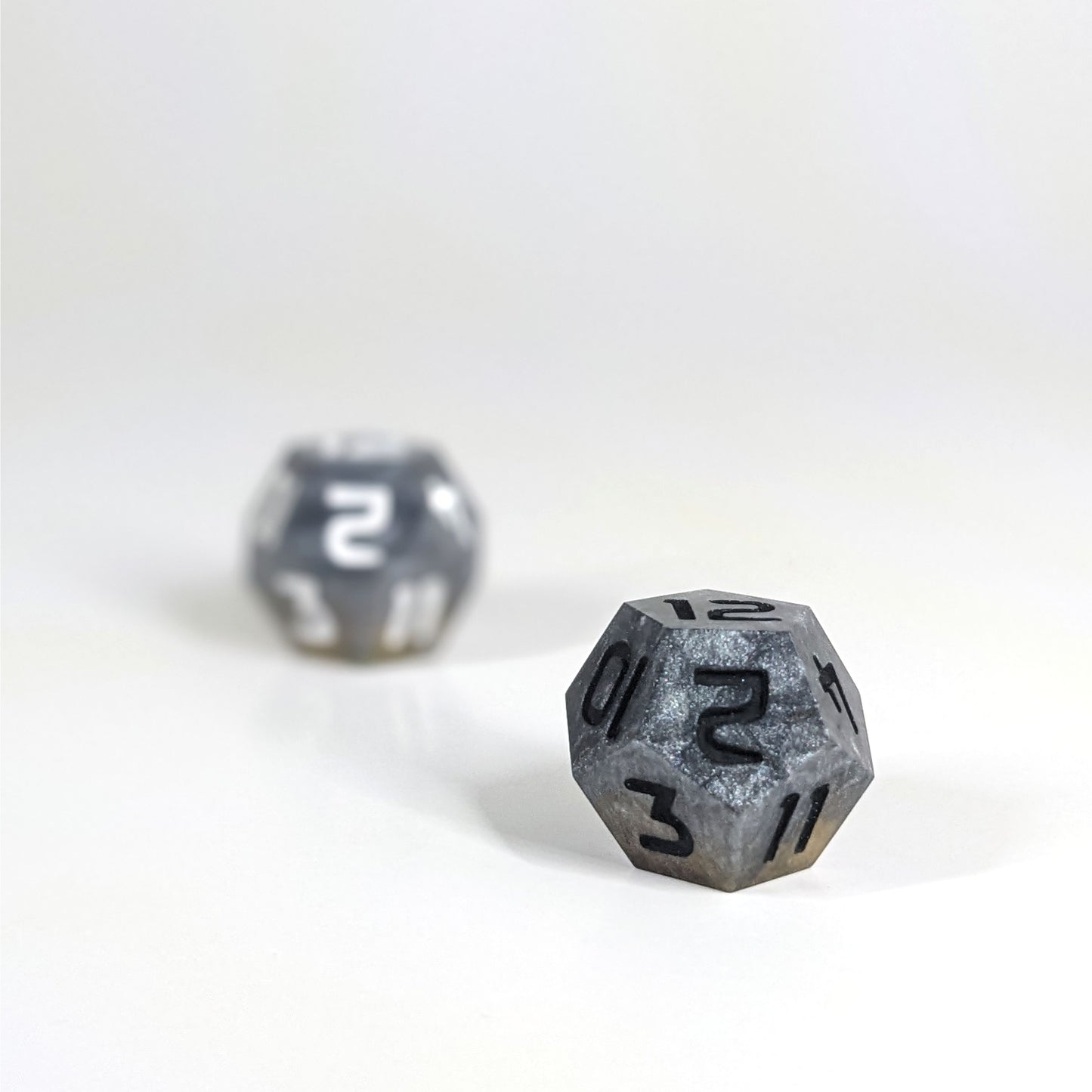 A low angle photo of two d12s. The dark silver and gold d12 with white numbers is on the left and is further from the camera and out of focus. The dark silver and gold d12 with black numbers is on the right and is closer to the camera and in sharp focus.