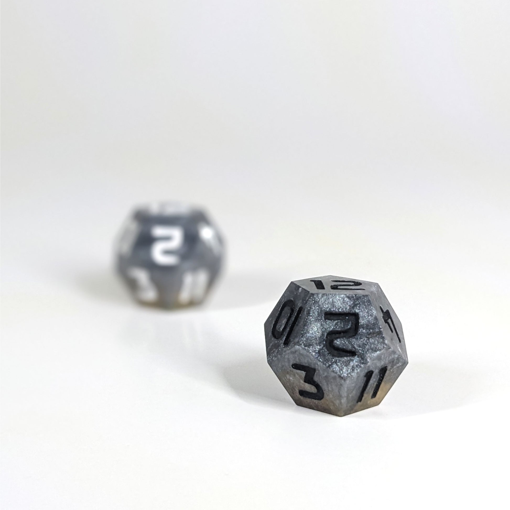 A low angle photo of two d12s. The dark silver and gold d12 with white numbers is on the left and is further from the camera and out of focus. The dark silver and gold d12 with black numbers is on the right and is closer to the camera and in sharp focus.