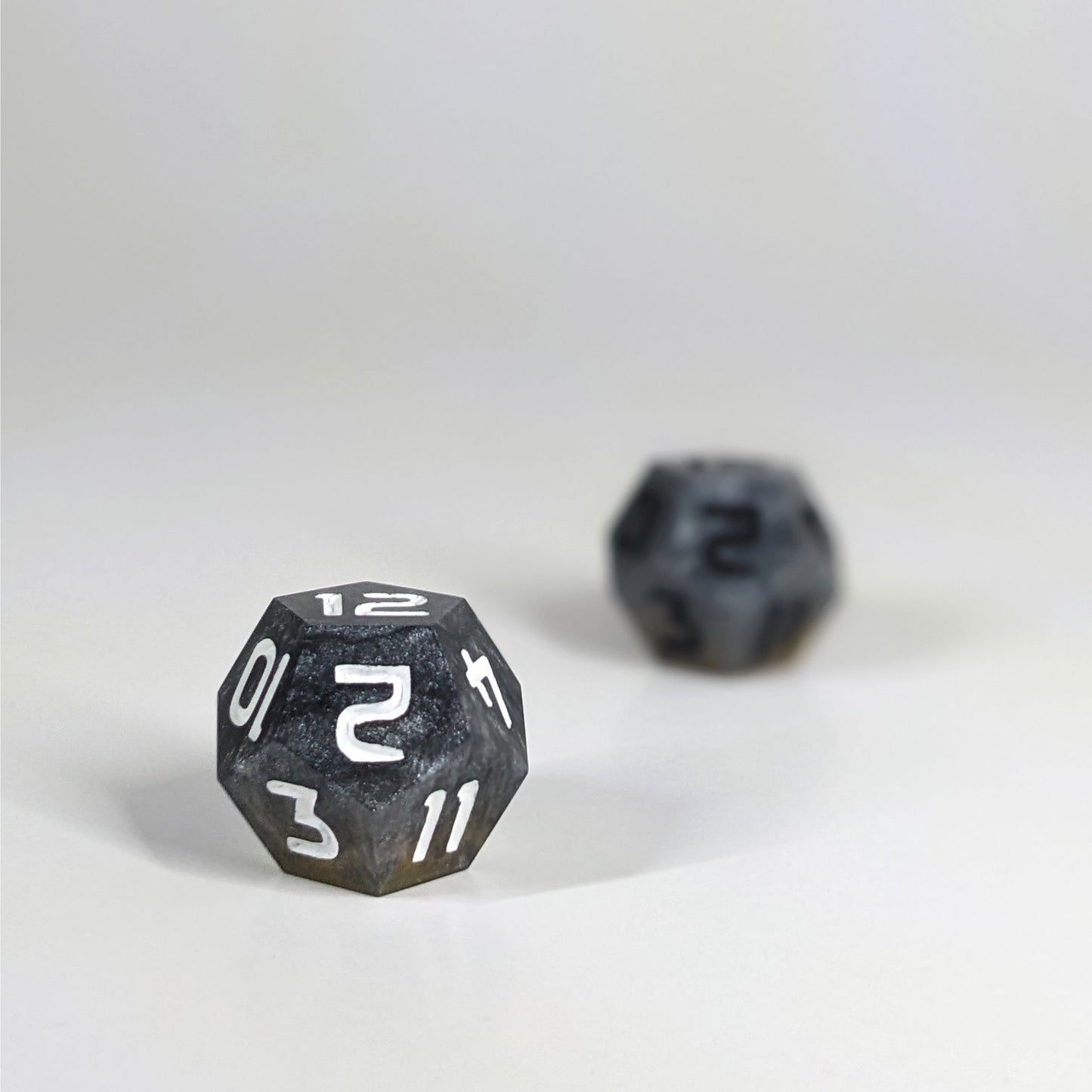 A low angle photo of two d12s. The dark silver and gold d12 with white numbers is on the left and is closer to the camera in sharp focus. The dark silver and gold d12 with black  numbers is on the right and is further from the camera and out of focus.