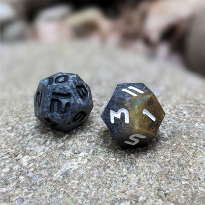 Two handmade d12s sit on a large rock. The dice are dark silver with a touch of gold, with one having black numbers and the other white numbers.