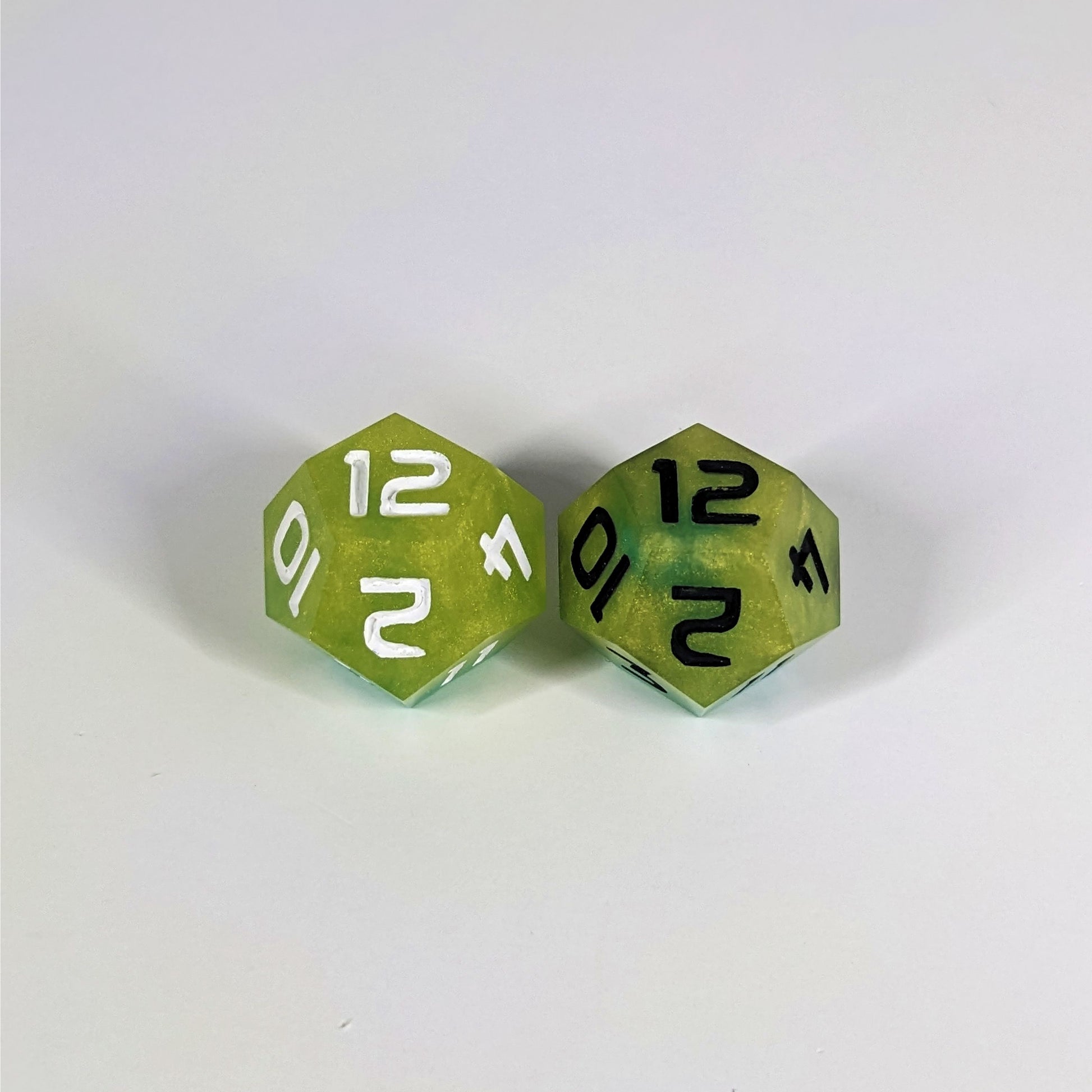 Two handmade d12s sit with the 12 faces up on a white background. The dice are yellow and teal. The die on the left has white numbers, the die on the right has black numbers.
