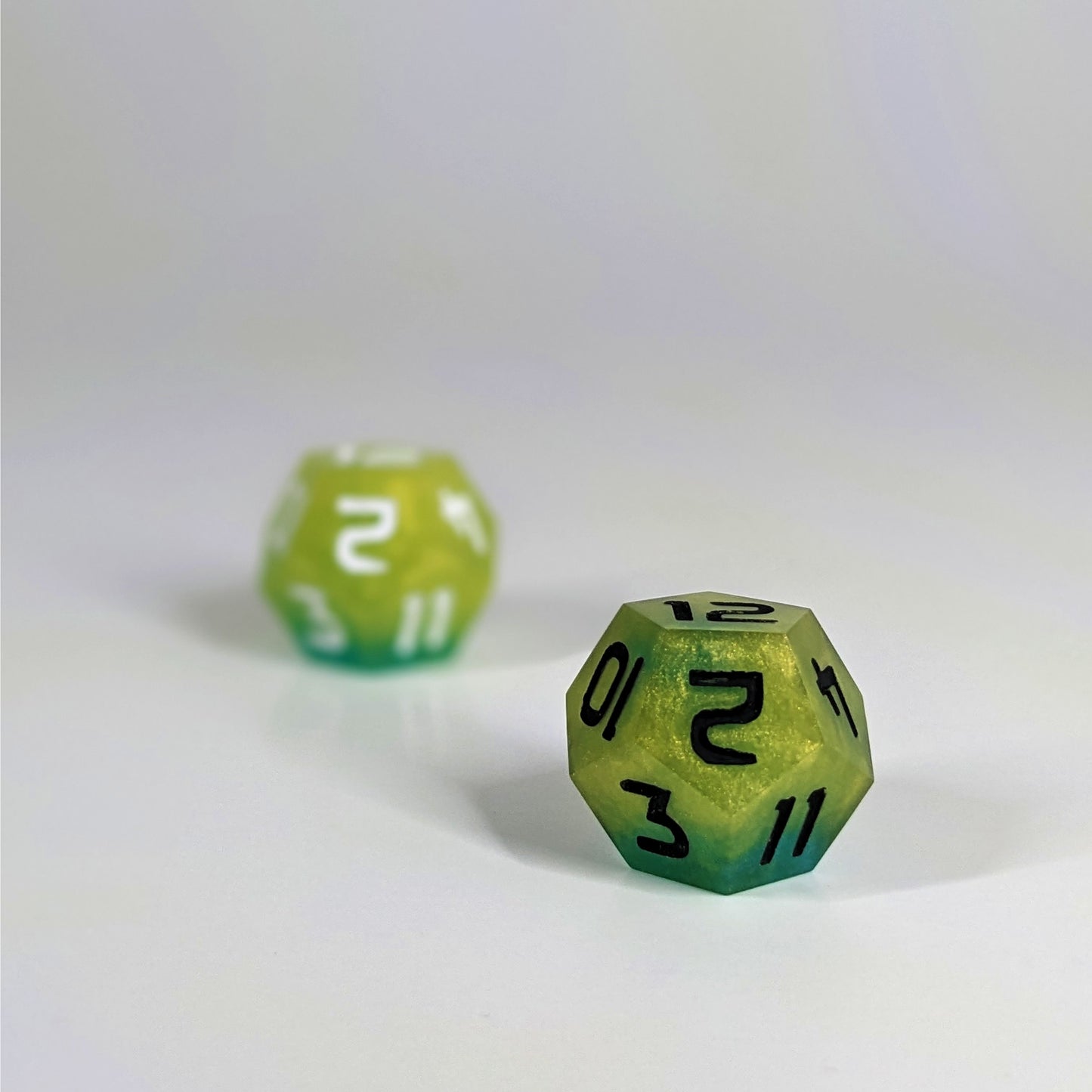 A low angle photo of two d12s. The yellow and teal d12 with white numbers is on the left and is further from the camera and out of focus. The yellow and teal d12 with black numbers is on the right and is closer to the camera and in sharp focus.