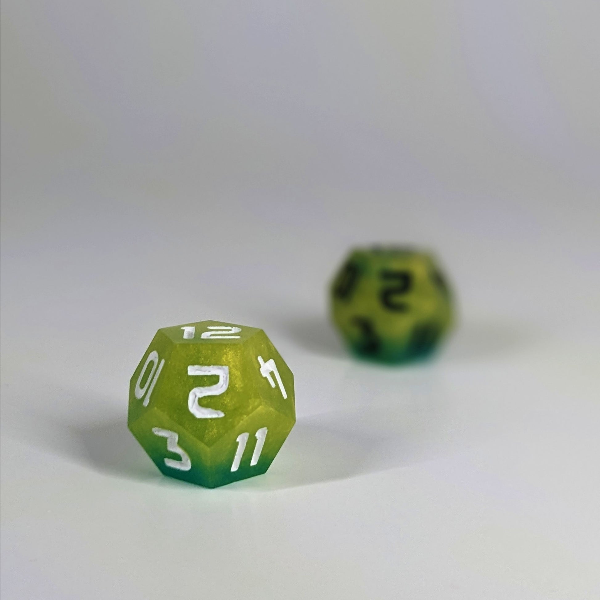 A low angle photo of two d12s. The yellow and teal d12 with white numbers is on the left and is closer to the camera in sharp focus. The yellow and teal d12 with black  numbers is on the right and is further from the camera and out of focus.