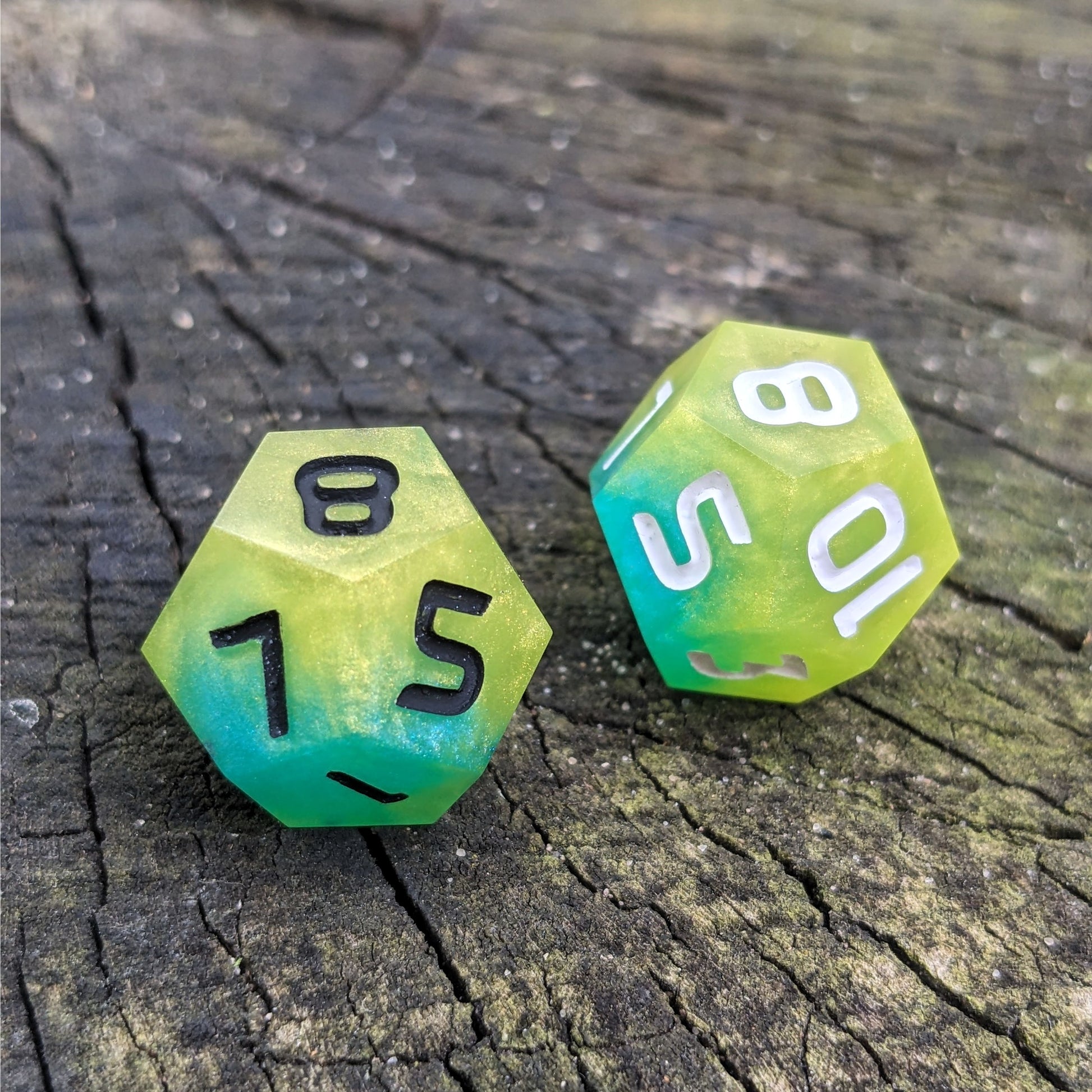 Two handmade d12s sit on a wood stump. The dice are yellow and teal, with one having black numbers and the other white numbers.