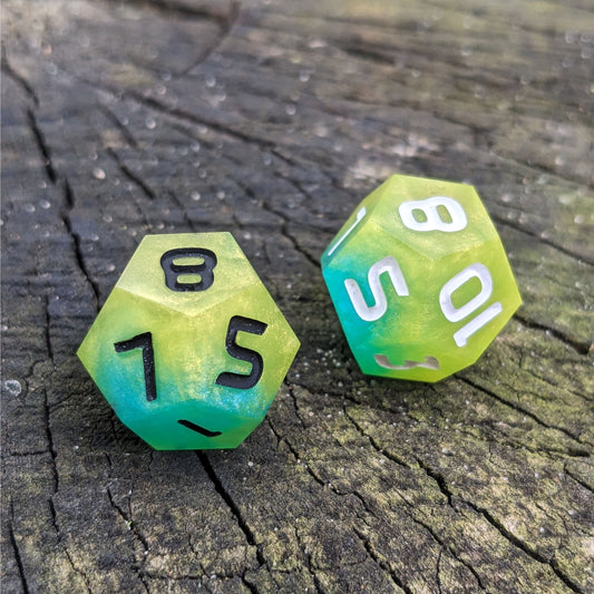 Two handmade d12s sit on a wood stump. The dice are yellow and teal, with one having black numbers and the other white numbers.