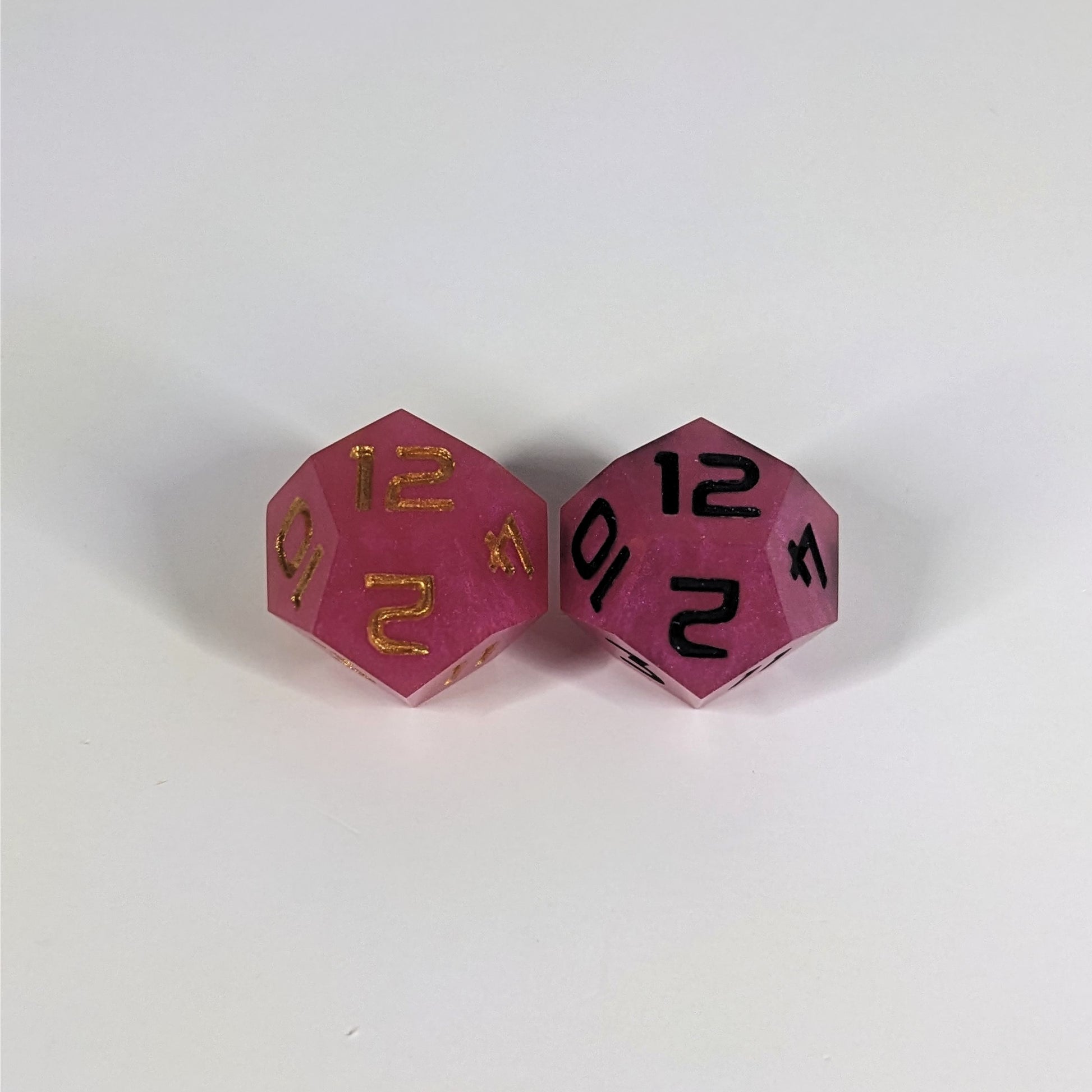Two handmade d12s sit with the 12 faces up on a white background. The dice are pink. The die on the left has gold numbers, the die on the right has black numbers.