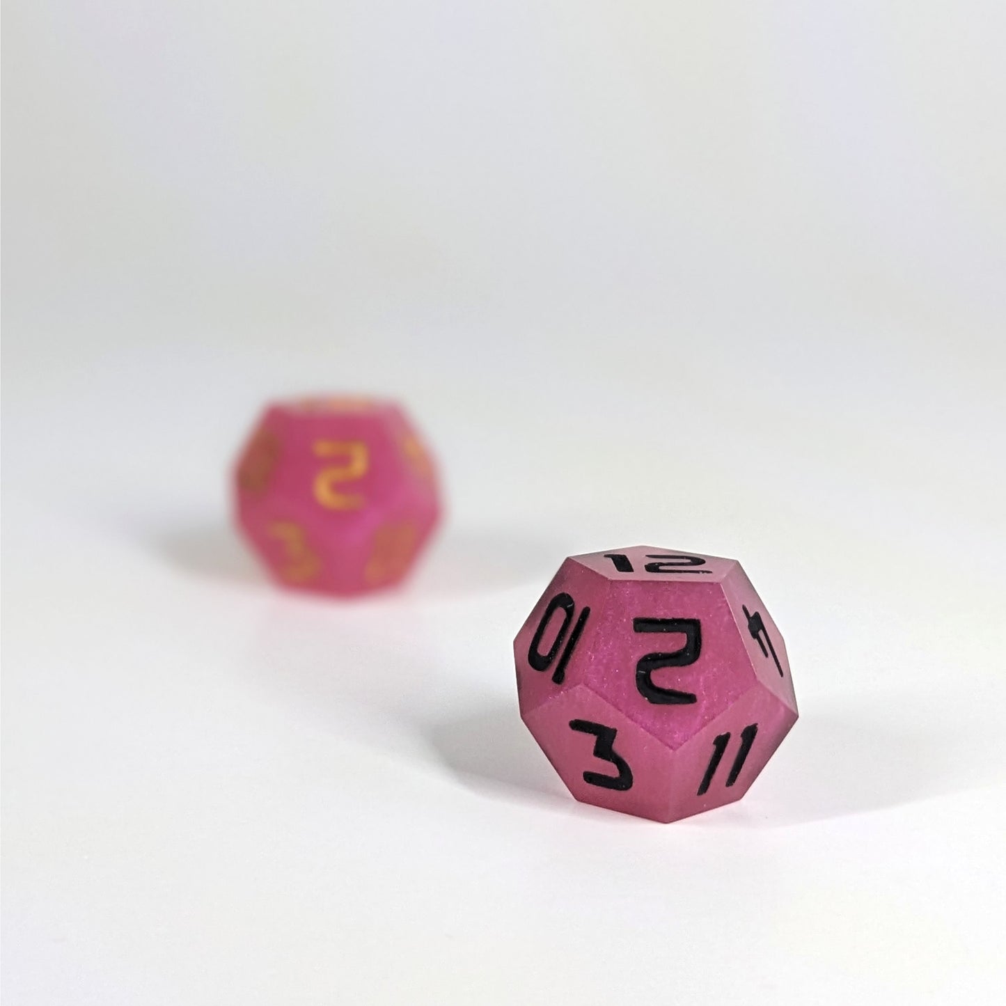 A low angle photo of two d12s. The pink d12 with gold numbers is on the left and is further from the camera and out of focus. The pink d12 with black numbers is on the right and is closer to the camera and in sharp focus.