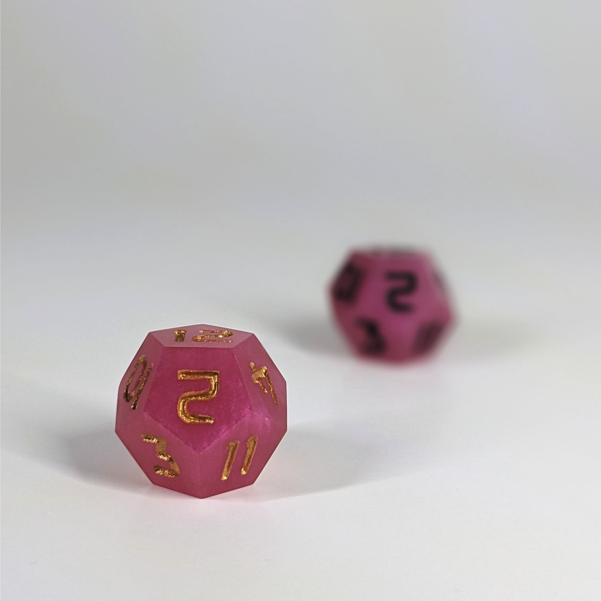 A low angle photo of two d12s. The pink d12 with gold numbers is on the left and is closer to the camera in sharp focus. The pink d12 with black  numbers is on the right and is further from the camera and out of focus.