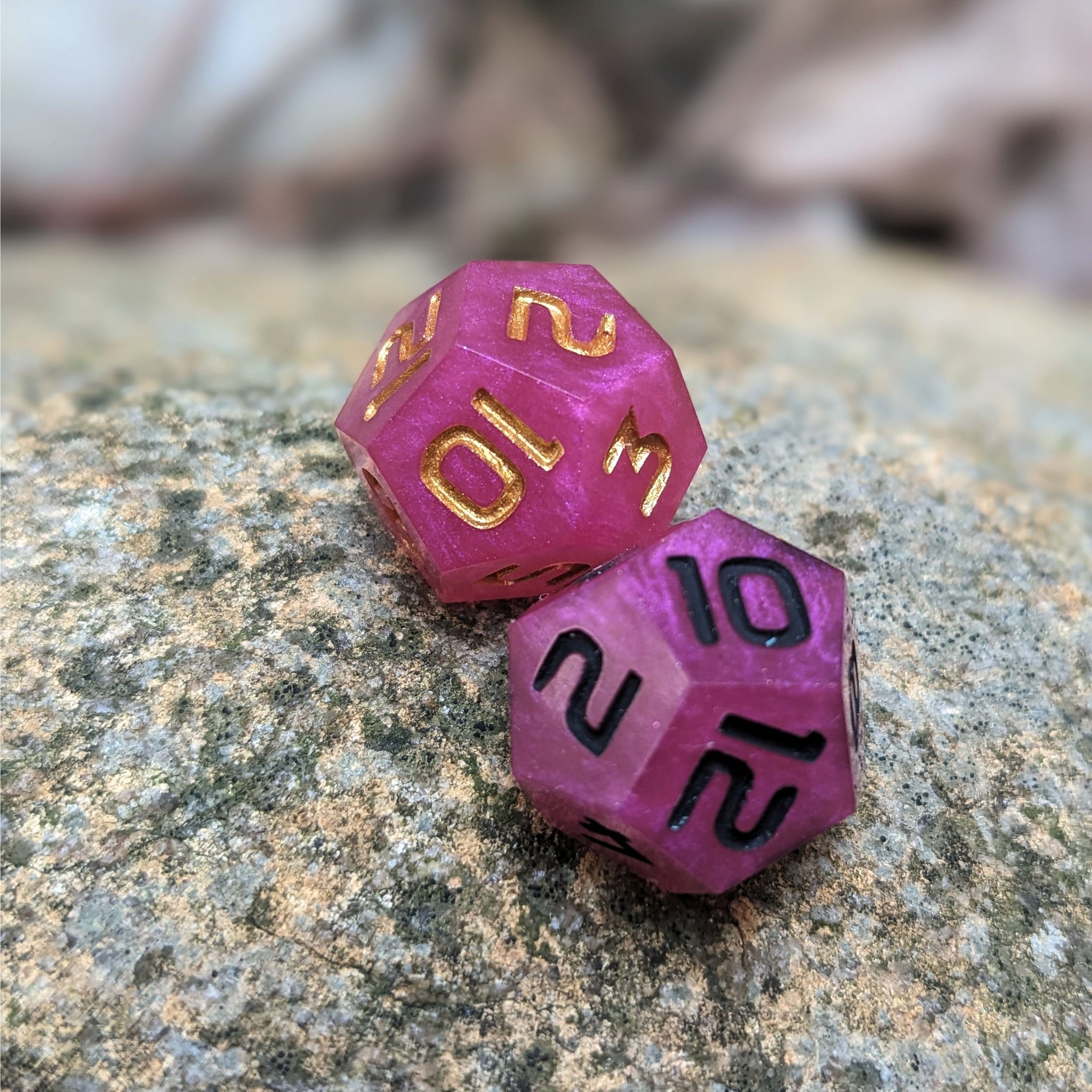 Two handmade d12s sit on a large rock. The dice are pink, with one having black numbers and the other gold numbers.