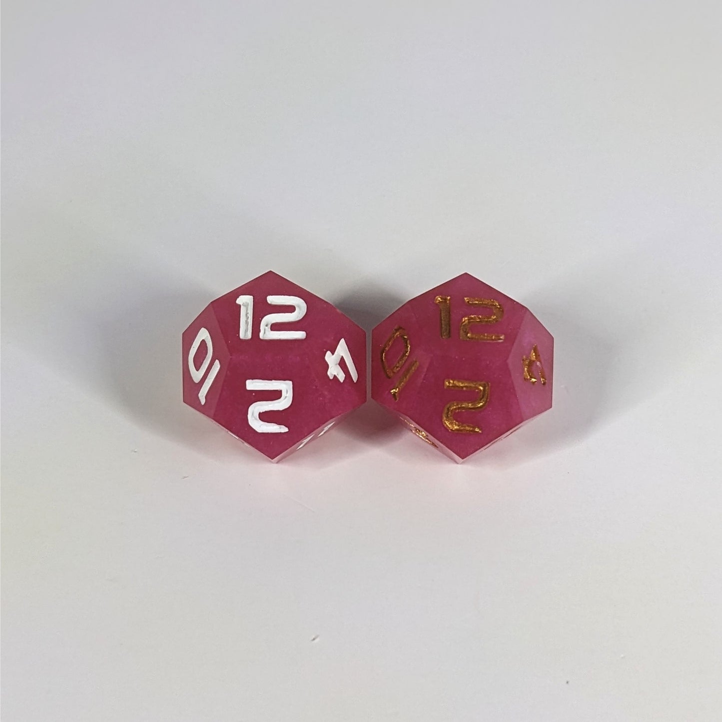 Two handmade d12s sit with the 12 faces up on a white background. The dice are pink. The die on the left has white numbers, the die on the right has gold numbers.