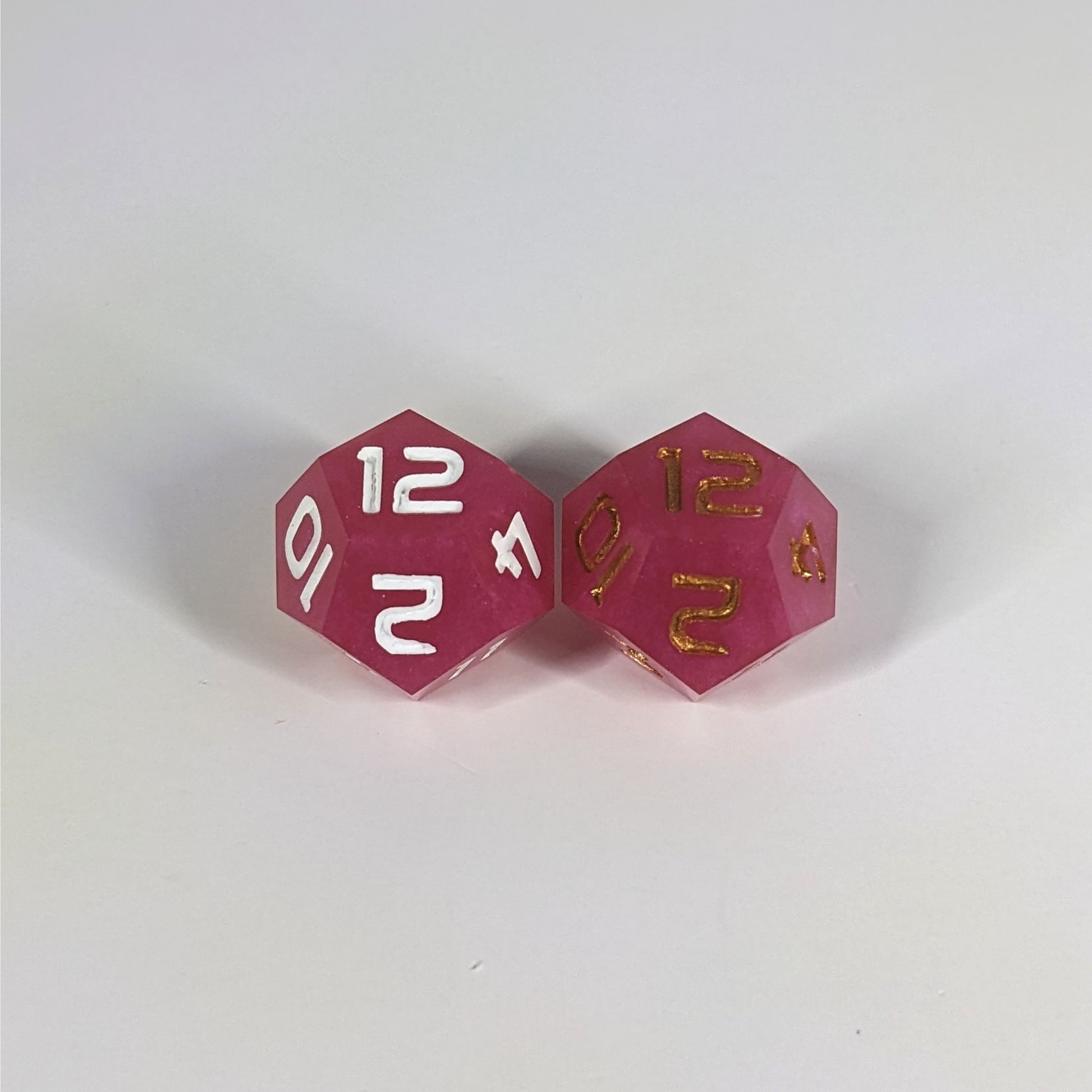 Two handmade d12s sit with the 12 faces up on a white background. The dice are pink. The die on the left has white numbers, the die on the right has gold numbers.