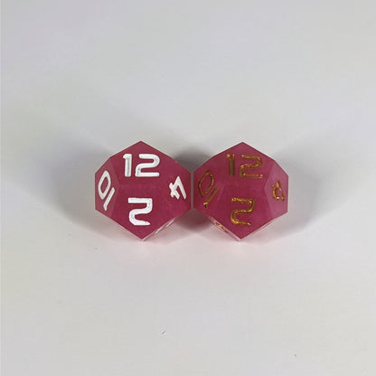 Two handmade d12s sit with the 12 faces up on a white background. The dice are pink. The die on the left has white numbers, the die on the right has gold numbers.