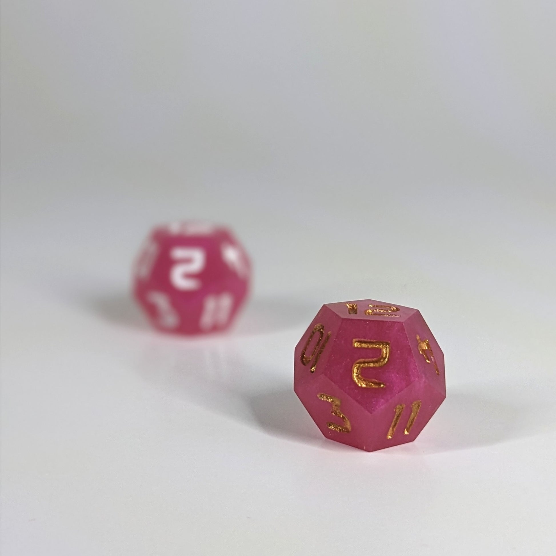 A low angle photo of two d12s. The pink d12 with white numbers is on the left and is further from the camera and out of focus. The pink d12 with gold numbers is on the right and is closer to the camera and in sharp focus.