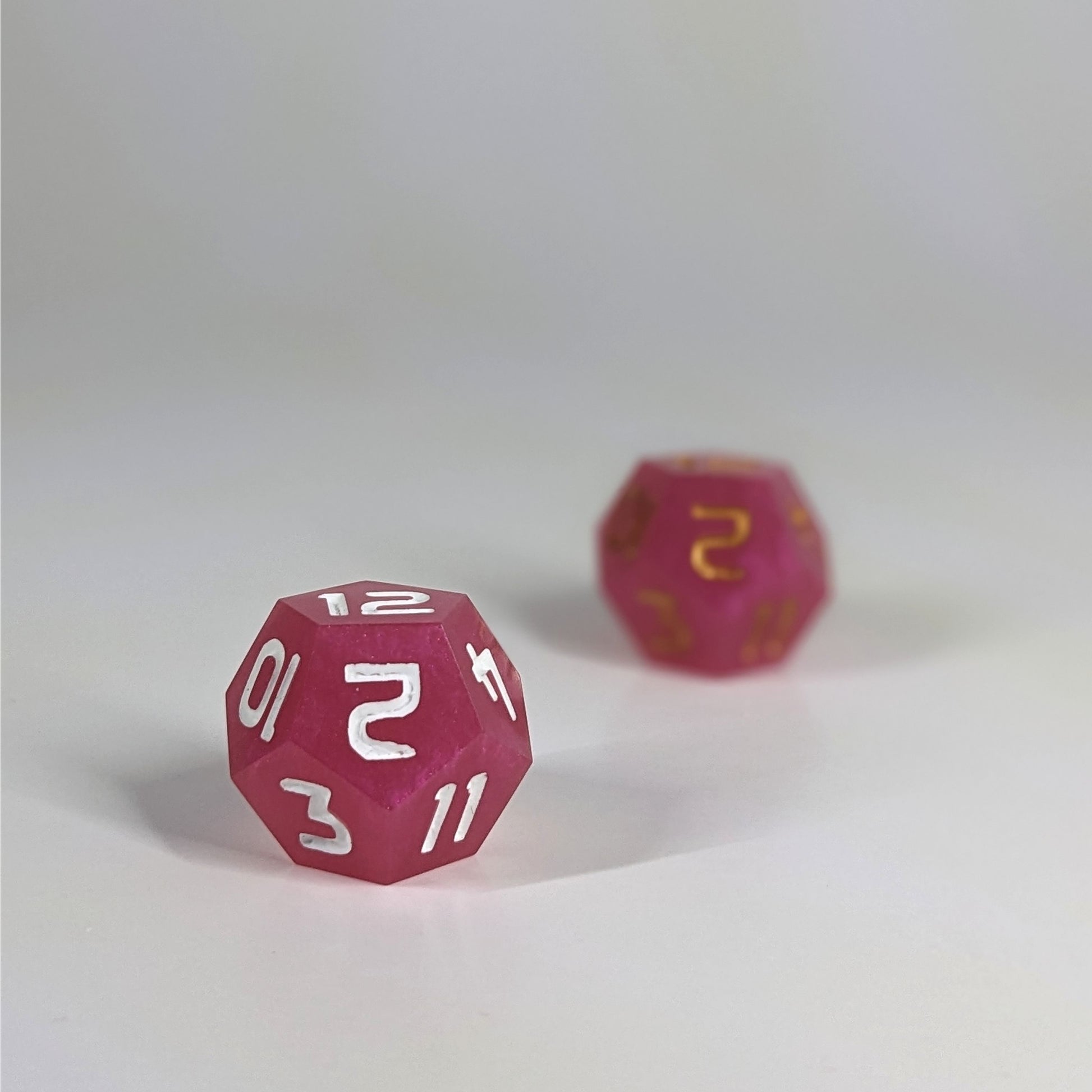 A low angle photo of two d12s. The pink d12 with white numbers is on the left and is closer to the camera in sharp focus. The pink d12 with gold numbers is on the right and is further from the camera and out of focus.