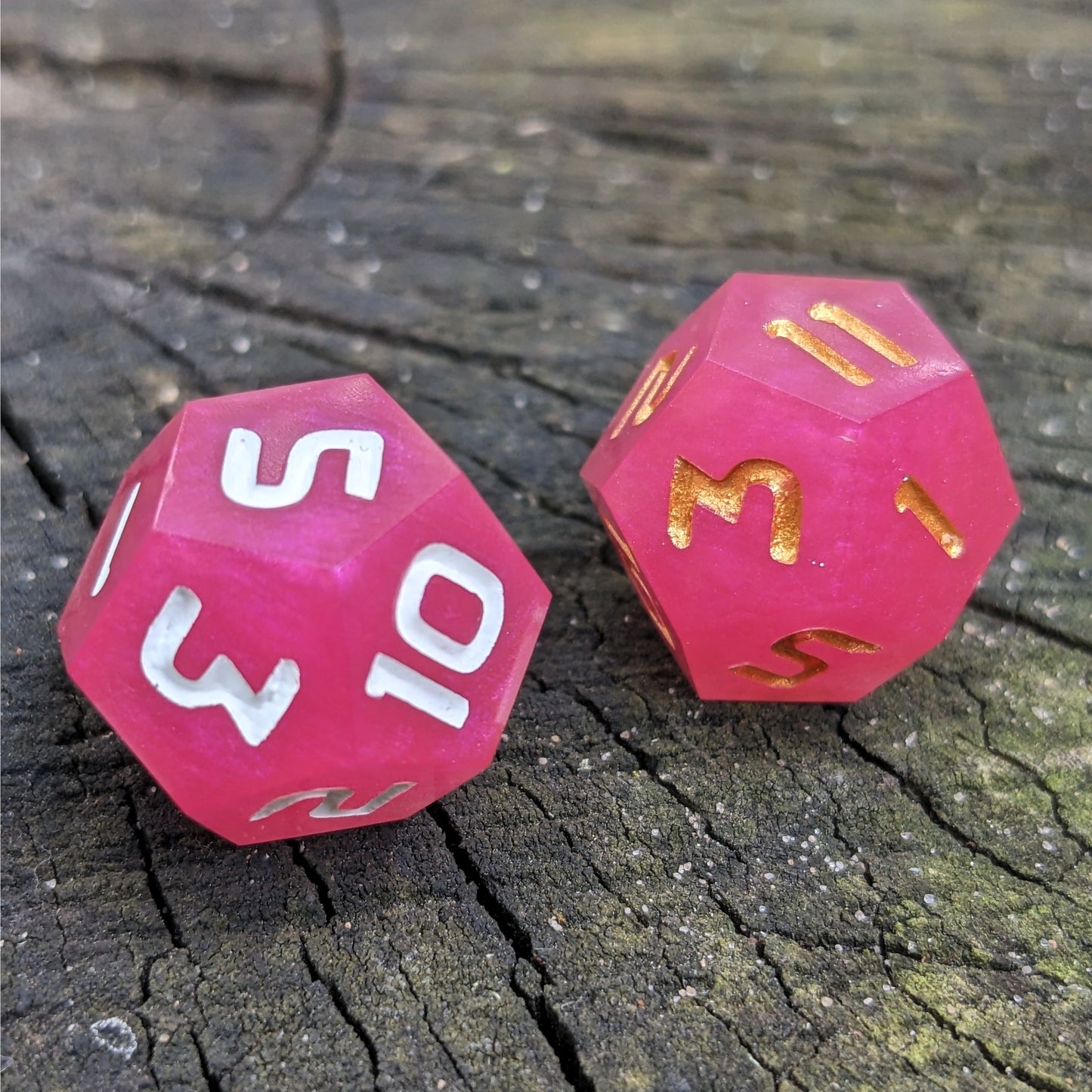 Two handmade d12s sit on a wood stump. The dice are pink, with one having white numbers and the other gold numbers.