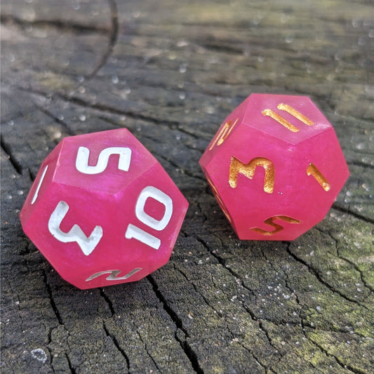 Two handmade d12s sit on a wood stump. The dice are pink, with one having white numbers and the other gold numbers.
