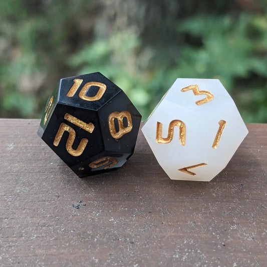 Two polyhedral dice, one black with gold numbers and letters, the other white with gold numbers and letters, on a wooden surface with a blurred green background.