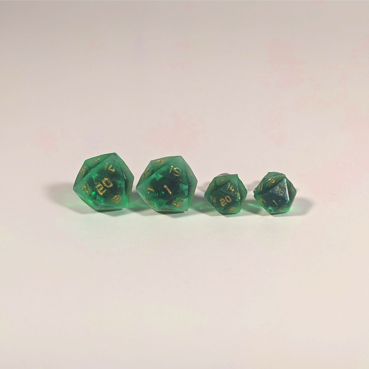 Two pairs of handmade d20 earrings with titanium hardware that are green with black flakies with gold numbers on a white background sit in a line with the larger pair on the left and the smaller pair on the right. The earrings on the left of each pair centers the 20 face and the earrings on the right of each pair centers the 1 face.