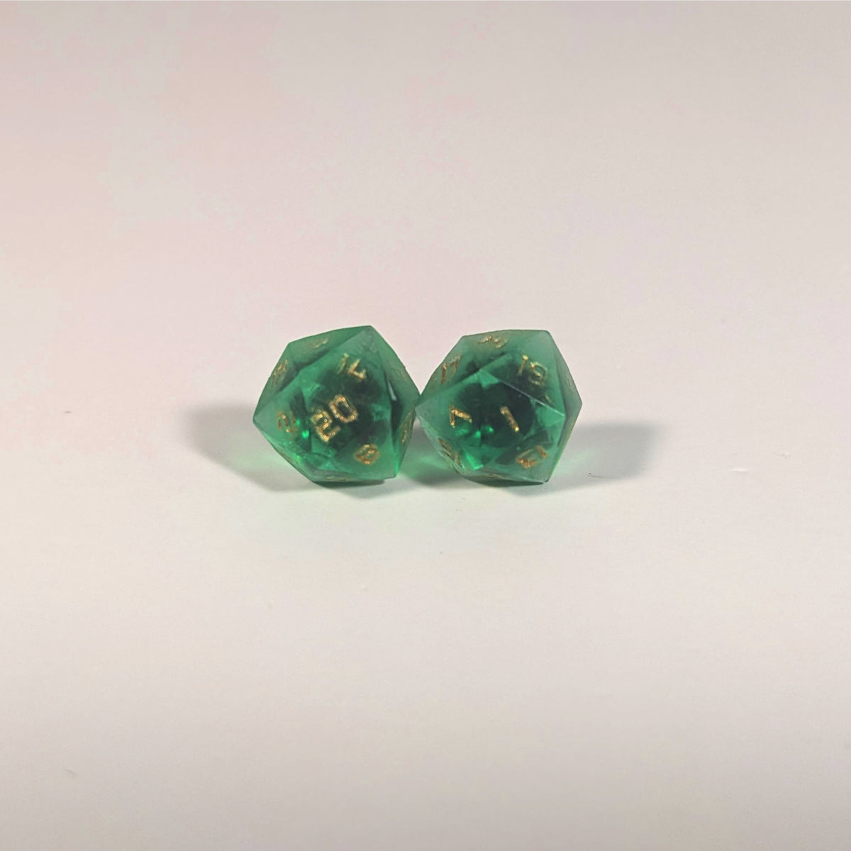 A single pair of the larger size handmade d20 earrings with titanium hardware that are green with black flakies with gold numbers on a white background. The earring on the left centers the 20 face and the earring on the right centers the 1 face.