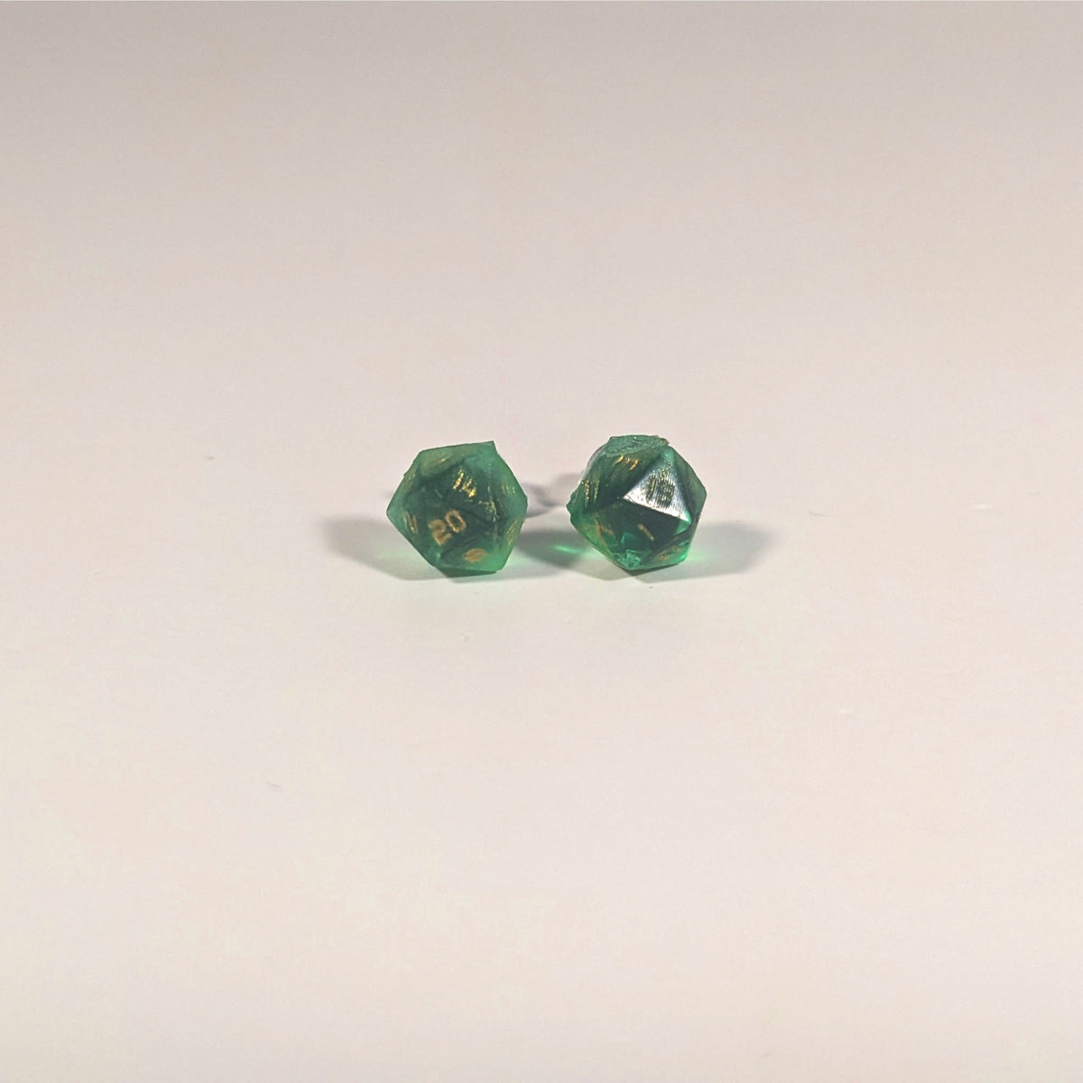 A single pair of the smaller size handmade d20 earrings with titanium hardware that are green with black flakies with gold numbers on a white background. The earring on the left centers the 20 face and the earring on the right centers the 1 face.