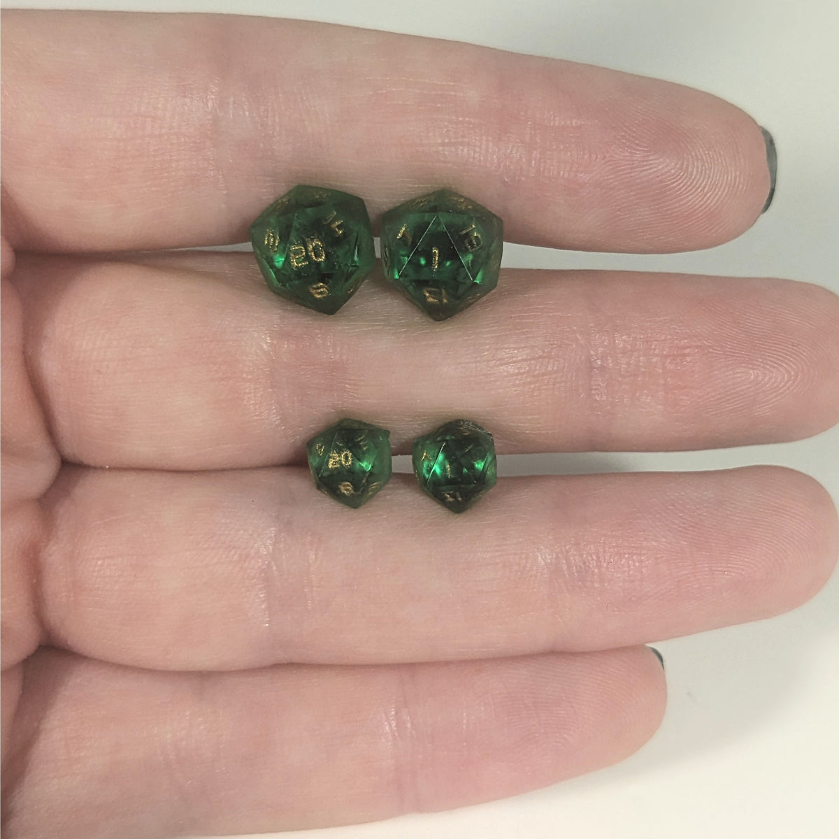 Two pairs of handmade d20 earrings with titanium hardware that are green with black flakies with gold numbers held between the fingers of a flat hand. The larger pair is between the pointer and middle finger, the smaller between the middle and ring finger. The earrings on the left of each pair centers the 20 face and the earrings on the right of each pair centers the 1 face.