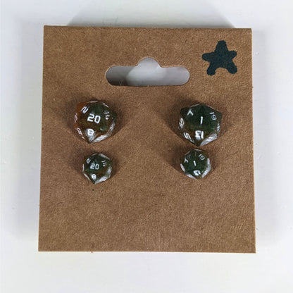 Two pairs of handmade d20 earrings with sterling silver hardware that are dark green and red gold with white numbers on a kraft brown earring card with a star in the top right corner. The earrings on the left of each pair centers the 20 face and the earrings on the right of each pair centers the 1 face.