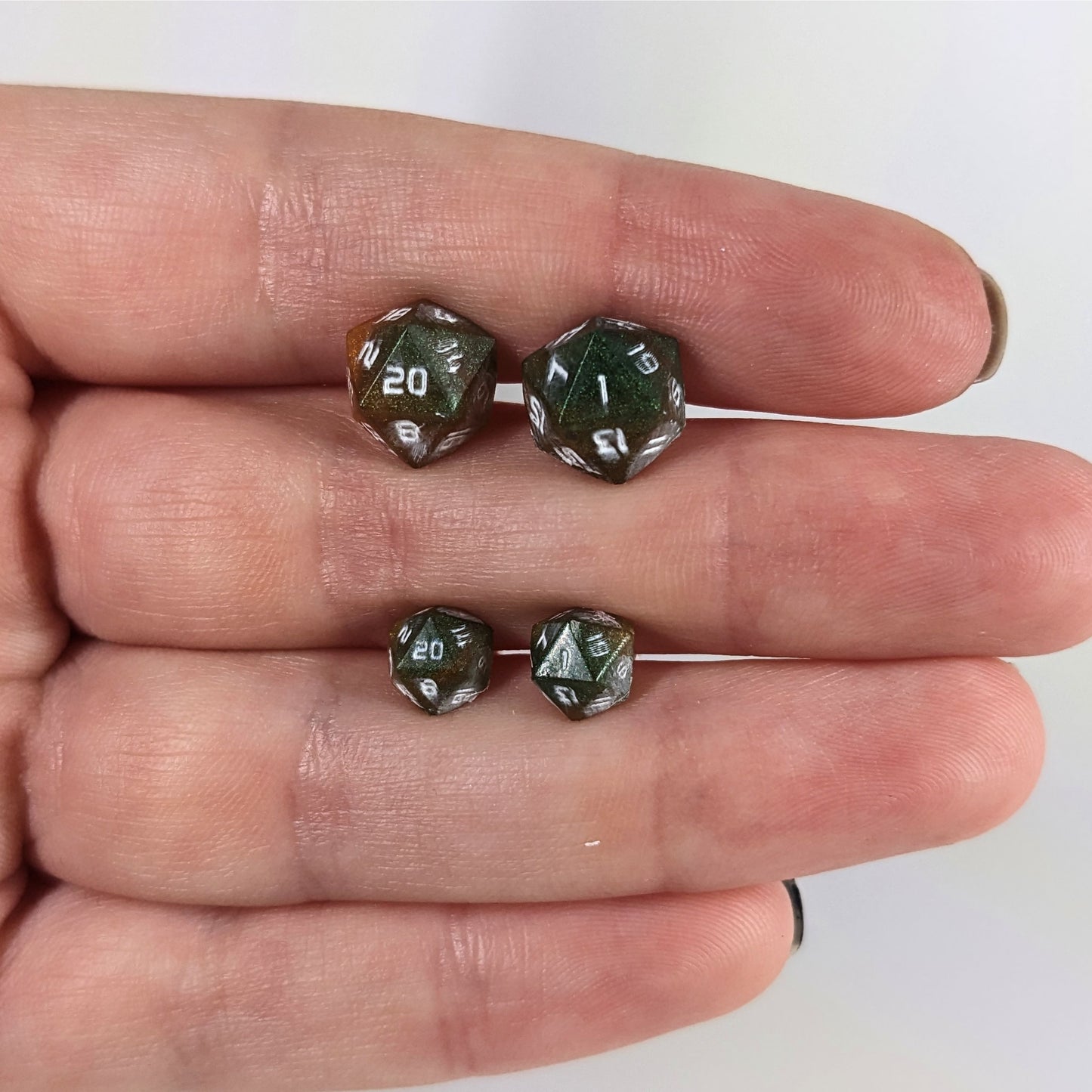 Two pairs of handmade d20 earrings with sterling silver hardware that are dark green and red gold with white numbers held between the fingers of a flat hand. The larger pair is between the pointer and middle finger, the smaller between the middle and ring finger. The earrings on the left of each pair centers the 20 face and the earrings on the right of each pair centers the 1 face.