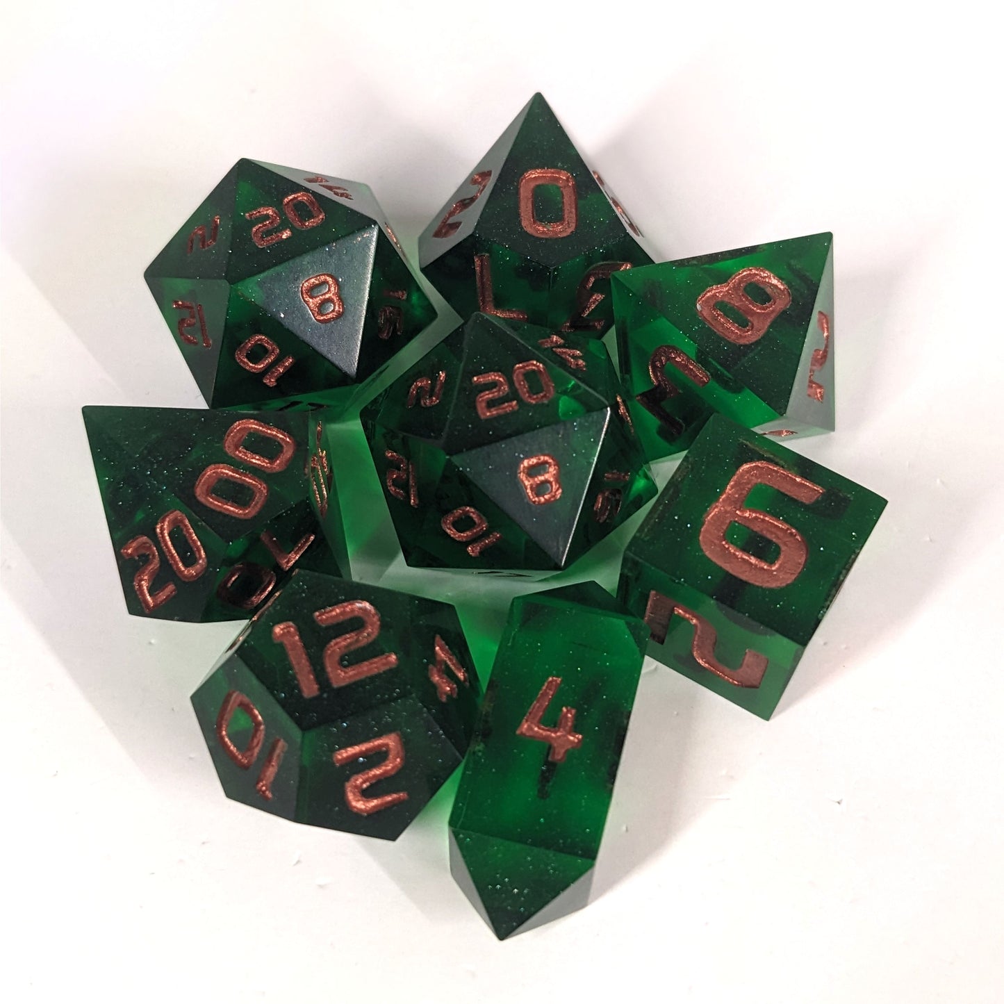 A complete set of 8 handmade artisan dice that are emerald green with copper numbers. The DnD dice set is arranged in a circle and features 2 D20s, one of which is in the center surrounded by the other 7 dice on a white background. The d4 is a unique, crystalline shape.