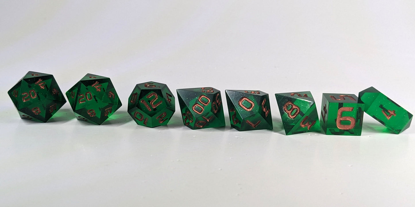 A straight on photo of a complete set of polyhedral dice that are emerald green with copper numbers arranged in a straight line. They are lined up with the 2 d20s on the left going down to the d4 on the right side. The highest value number of each die is facing forward, and the crystalline d4 is leaning against the d6.