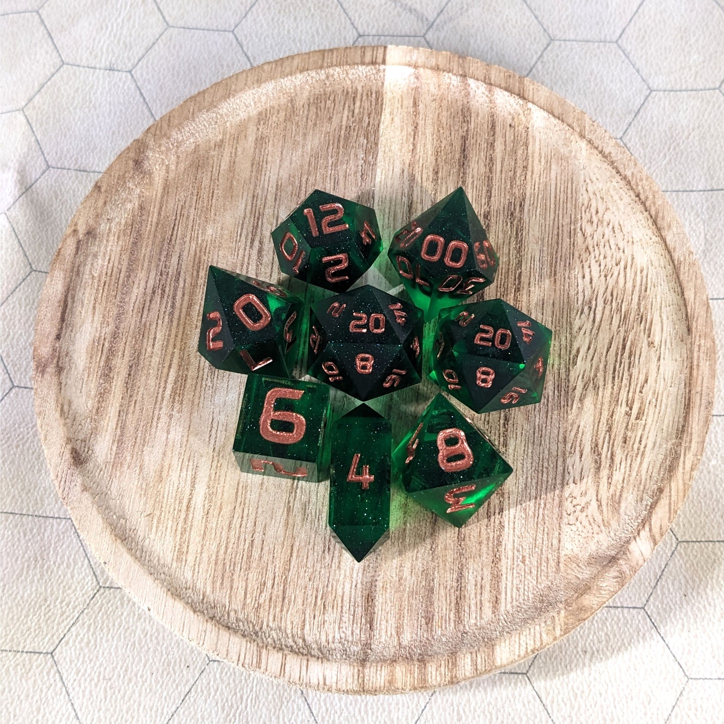 A complete set of 8 handmade artisan dice that are emerald green with copper numbers. The DnD dice set is on a wooded circle and the dice are arranged in a circle and features 2 D20s, one of which is in the center surrounded by the other 7 dice. The d4 is unique with a crystalline shape.