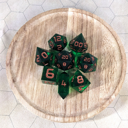A complete set of 8 handmade artisan dice that are emerald green with copper numbers. The DnD dice set is on a wooded circle and the dice are arranged in a circle and features 2 D20s, one of which is in the center surrounded by the other 7 dice. The d4 is unique with a crystalline shape.