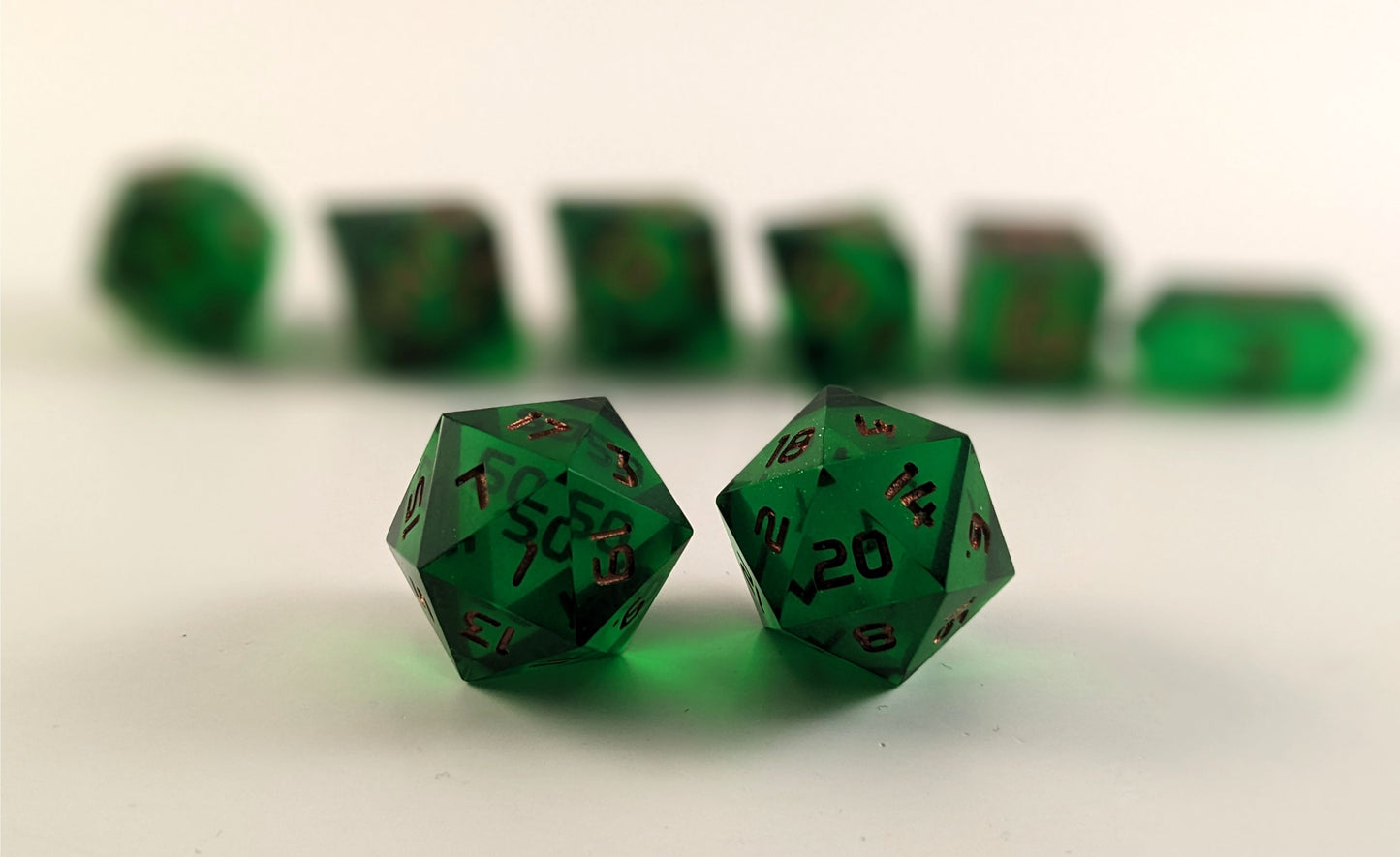A straight on photo of a standard set of DnD dice that are emerald green with copper numbers focusing on 2 d20s in the center. The d20 on the left has the 1 face centered and the d20 on the right has the 20 face centered. The other 6 dice are lined up in the background but are out of focus, while the d20s are in sharp focus.
