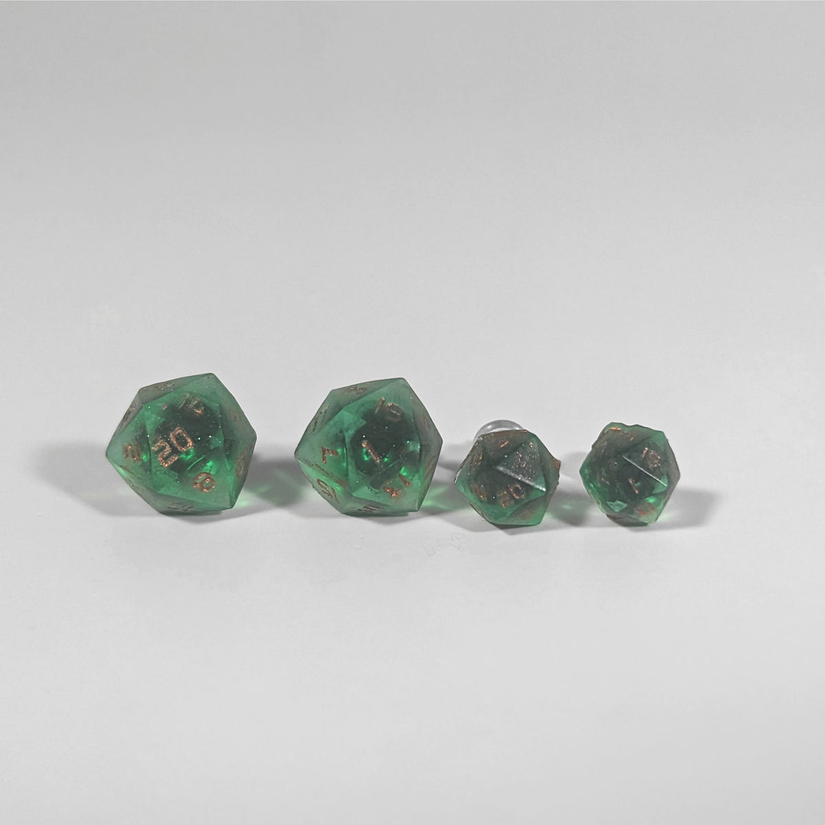 Two pairs of handmade d20 earrings with titanium hardware that are emerald green with copper numbers on a white background sit in a line with the larger pair on the left and the smaller pair on the right. The earrings on the left of each pair centers the 20 face and the earrings on the right of each pair centers the 1 face.