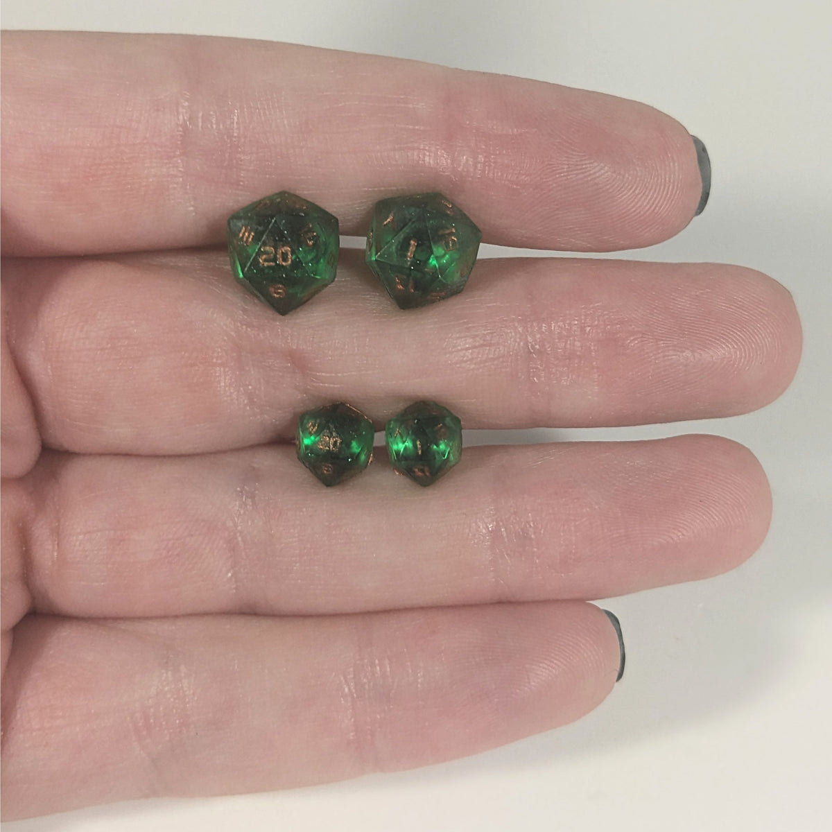Two pairs of handmade d20 earrings with titanium hardware that are emerald green with copper numbers held between the fingers of a flat hand. The larger pair is between the pointer and middle finger, the smaller between the middle and ring finger. The earrings on the left of each pair centers the 20 face and the earrings on the right of each pair centers the 1 face.