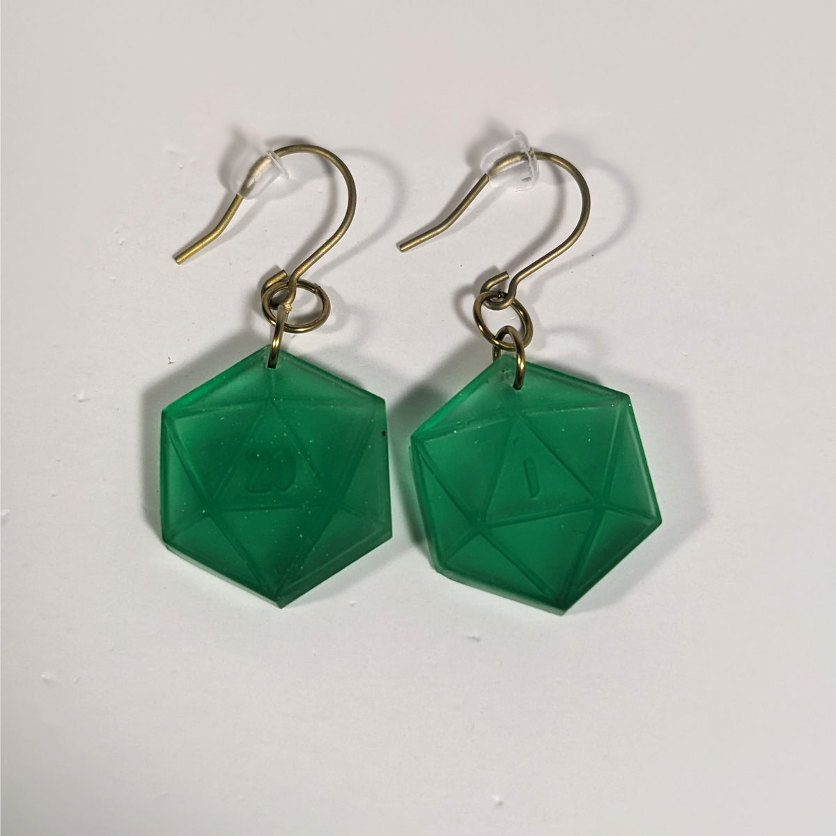 A pair of emerald green dangly, flat d20 earrings with gold color titanium and niobium hardware and bell shaped rubber backs on a white background. The earring on the left has a 20 on it and the earring on the right has a 1 on it, both in a fantasy style font.