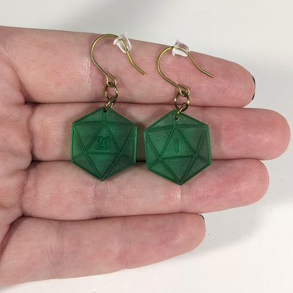 A pair of emerald green dangly, flat d20 earrings with gold color titanium and niobium hardware with bell shaped rubber backs held on on a hand on a white background. The earring on the left has a 20 on it and the earring on the right has a 1 on it, both in a fantasy style font.