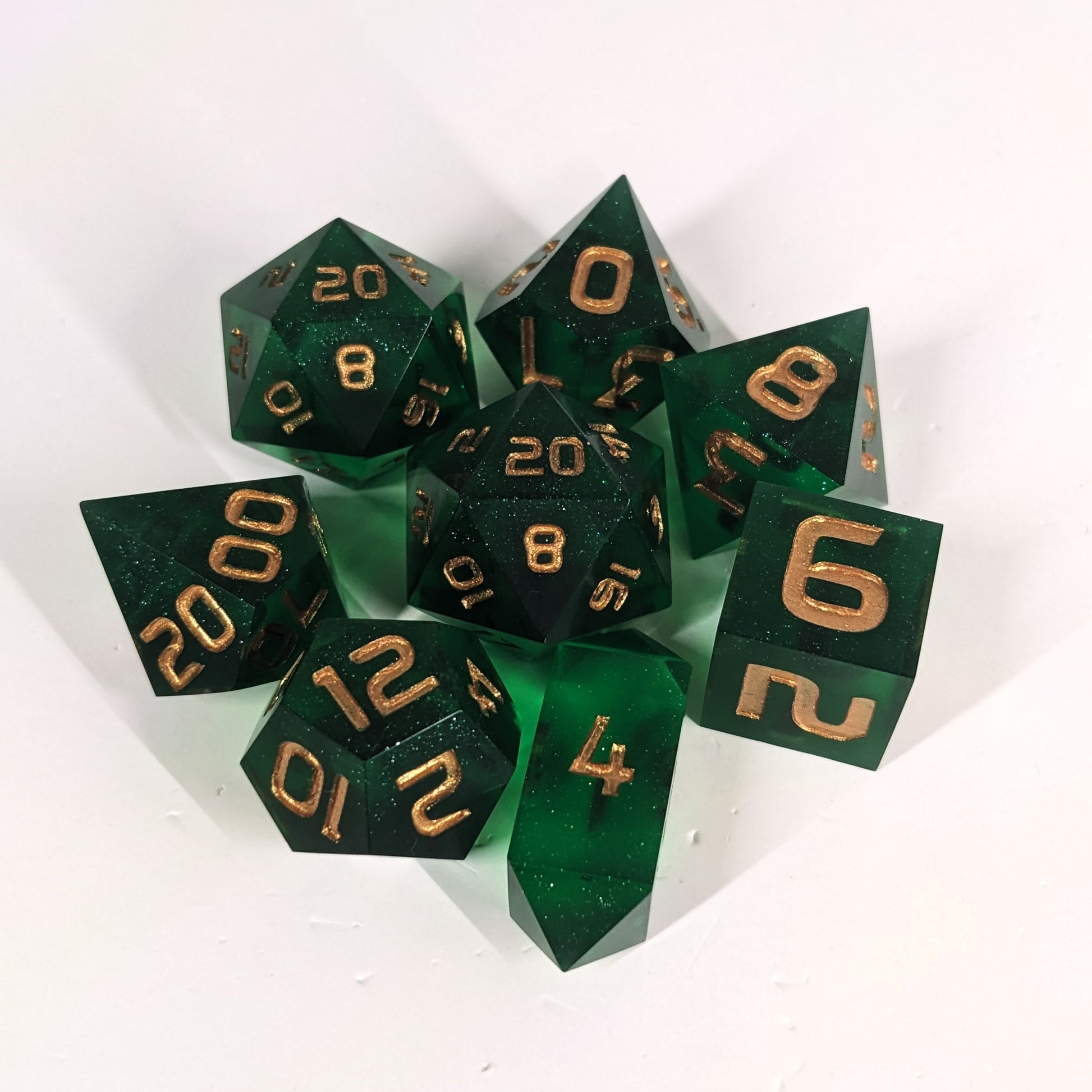 A complete set of 8 handmade artisan dice that are emerald green with gold numbers. The DnD dice set is arranged in a circle and features 2 D20s, one of which is in the center surrounded by the other 7 dice on a white background. The d4 is a unique, crystalline shape.