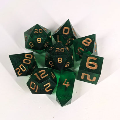 A complete set of 8 handmade artisan dice that are emerald green with gold numbers. The DnD dice set is arranged in a circle and features 2 D20s, one of which is in the center surrounded by the other 7 dice on a white background. The d4 is a unique, crystalline shape.