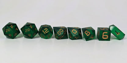 A straight on photo of a complete set of polyhedral dice that are emerald green with gold numbers arranged in a straight line. They are lined up with the 2 d20s on the left going down to the d4 on the right side. The highest value number of each die is facing forward, and the crystalline d4 is leaning against the d6.