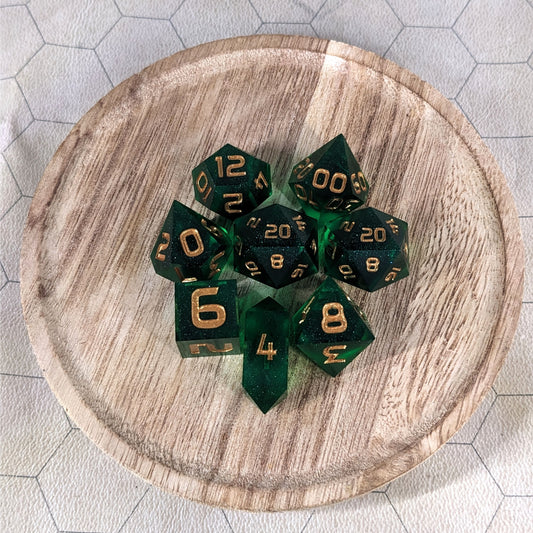 A complete set of 8 handmade artisan dice that are emerald green with gold numbers. The DnD dice set is on a wooded circle and the dice are arranged in a circle and features 2 D20s, one of which is in the center surrounded by the other 7 dice. The d4 is unique with a crystalline shape.