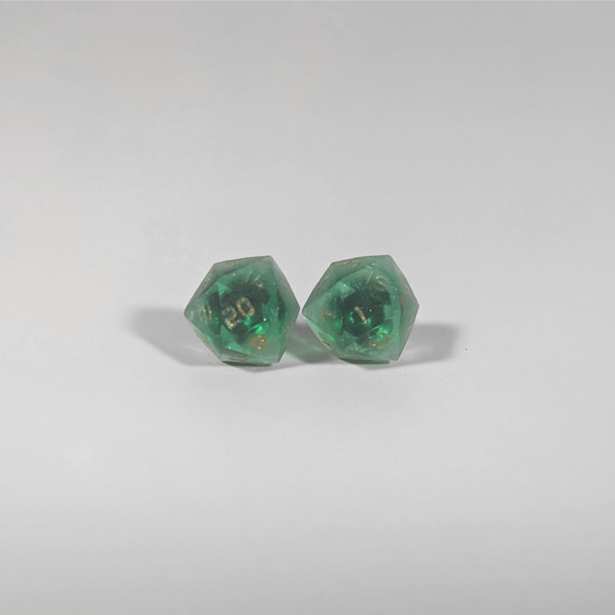 A single pair of the larger size handmade d20 earrings with titanium hardware that are emerald green with gold numbers on a white background. The earring on the left centers the 20 face and the earring on the right centers the 1 face.