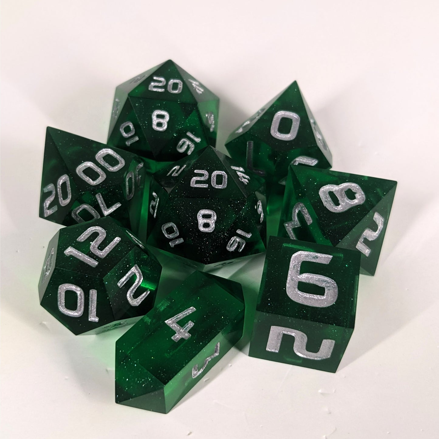 A complete set of 8 handmade artisan dice that are emerald green with silver numbers. The DnD dice set is arranged in a circle and features 2 D20s, one of which is in the center surrounded by the other 7 dice on a white background. The d4 is a unique, crystalline shape.
