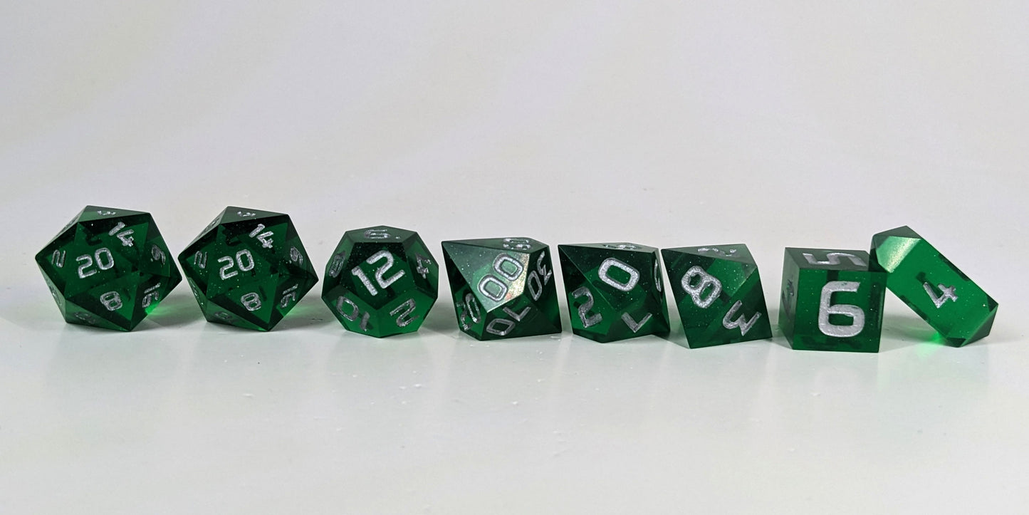 A straight on photo of a complete set of polyhedral dice that are emerald green with silver numbers arranged in a straight line. They are lined up with the 2 d20s on the left going down to the d4 on the right side. The highest value number of each die is facing forward, and the crystalline d4 is leaning against the d6.