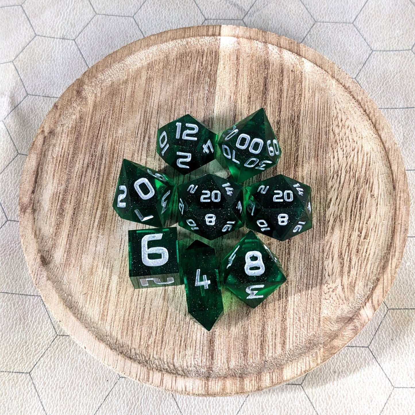 A complete set of 8 handmade artisan dice that are emerald green with silver numbers. The DnD dice set is on a wooded circle and the dice are arranged in a circle and features 2 D20s, one of which is in the center surrounded by the other 7 dice. The d4 is unique with a crystalline shape.