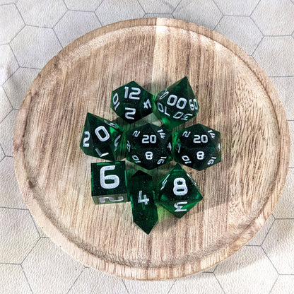 A complete set of 8 handmade artisan dice that are emerald green with silver numbers. The DnD dice set is on a wooded circle and the dice are arranged in a circle and features 2 D20s, one of which is in the center surrounded by the other 7 dice. The d4 is unique with a crystalline shape.