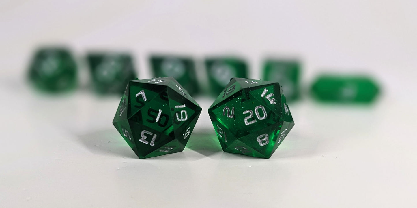 A straight on photo of a standard set of DnD dice that are emerald green with silver numbers focusing on 2 d20s in the center. The d20 on the left has the 1 face centered and the d20 on the right has the 20 face centered. The other 6 dice are lined up in the background but are out of focus, while the d20s are in sharp focus.