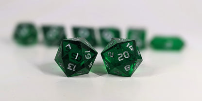 A straight on photo of a standard set of DnD dice that are emerald green with silver numbers focusing on 2 d20s in the center. The d20 on the left has the 1 face centered and the d20 on the right has the 20 face centered. The other 6 dice are lined up in the background but are out of focus, while the d20s are in sharp focus.