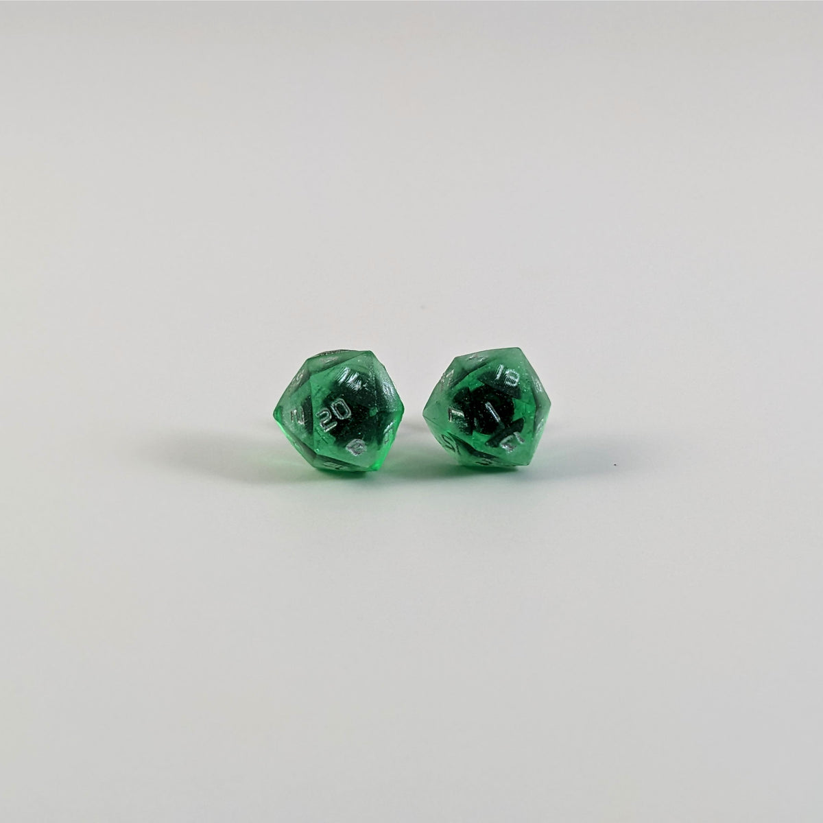 A single pair of the larger size handmade d20 earrings with titanium hardware that are emerald green with silver numbers on a white background. The earring on the left centers the 20 face and the earring on the right centers the 1 face.